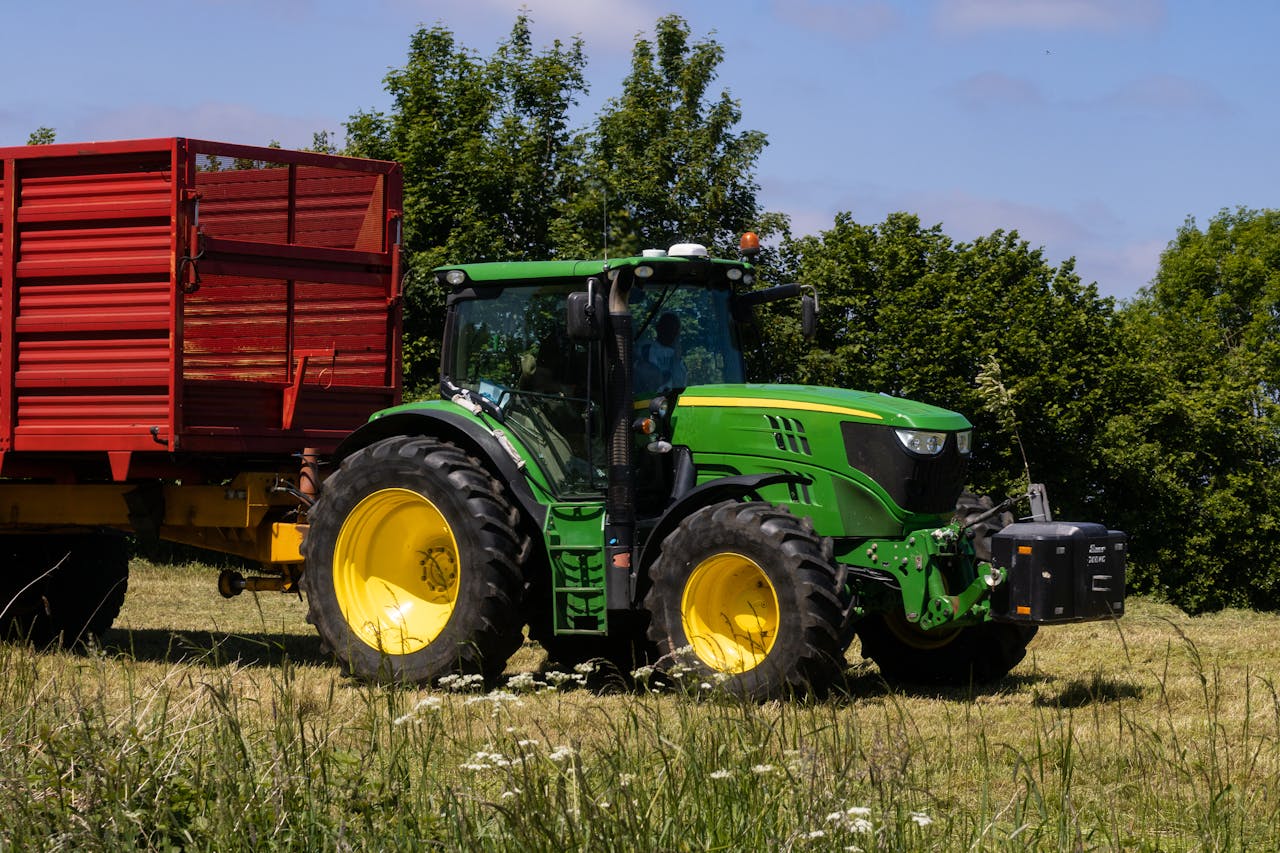 our-story A green tractor with a yellow wheel pulls a red trailer in a sunny countryside field.